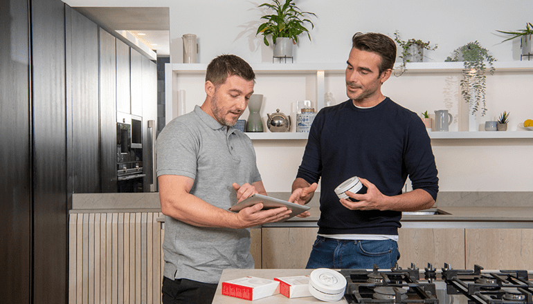 Two men in a kitchen looking at a smoke or CO alarm