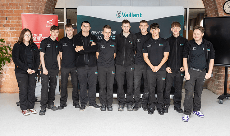 A group of low-carbon heating technician apprentices stand in a line in front of a sign showing Vaillant's logo at Derby College