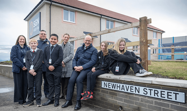 First Minister John Swinney and SGN CEO Simon Kilonback with members of community leaning on a wall outside a hydrogen demonstration house