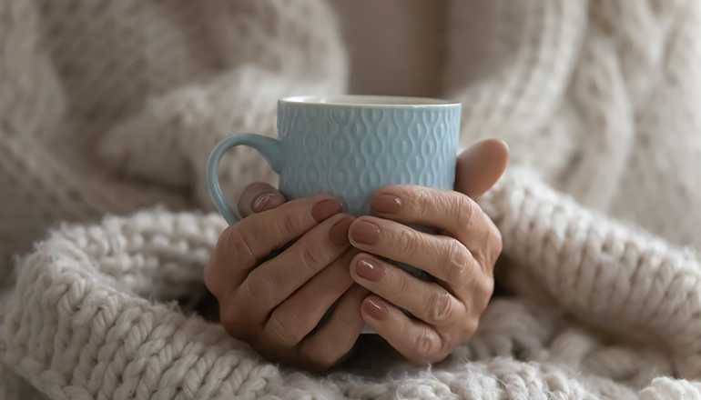 A woman's hands wrapped around a mug containing a warm drink, indicating being cold