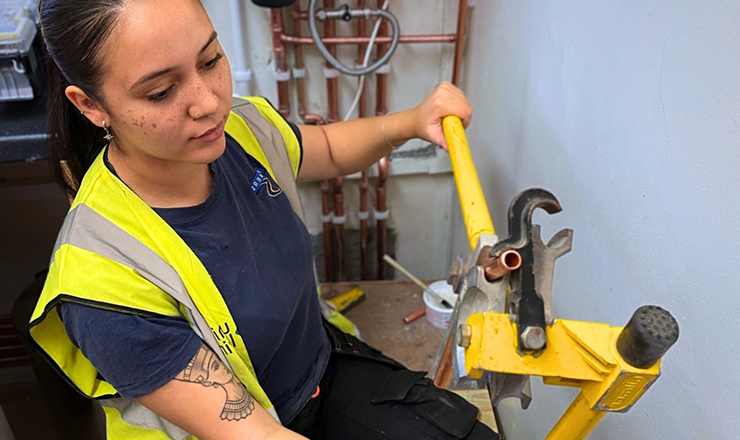 A young female plumbing apprentice wearing a high-vis jacket sitting a bench bending copper pipe